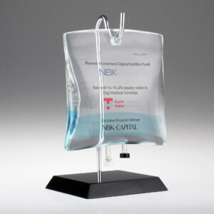 Transparent glass deal toys on a metal base with engraved text about a financial transaction involving the Kuwait investment opportunities fund and elaj medical services, dated February 2017.