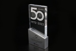 A transparent glass trophy commemorating 50 years, from 1972 to 2022, displayed against a black background. the recognition award has an inscription and logos.