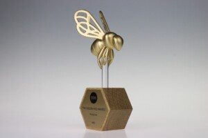 Golden recognition award resembling a bee mounted on a hexagonal base with the inscription "bbn the golden bee awards - honoring best - 2022". the trophy is set against a grey gradient background.