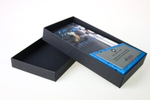 A black commemorative box with an open lid, containing a framed photo and a metallic plaque with engraved text on a blue background.