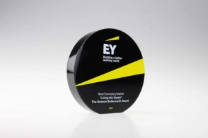 A sleek black circular recognition award with ey logo and yellow diagonal stripes, text reading "building a better working world, most exemplary market 'living the dream' award, the graeme button award, uk.