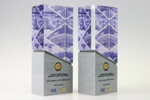 Two identical custom recognition awards featuring a base of speckled grey material with a printed upper section depicting construction equipment. each is labeled with event and sponsor details.