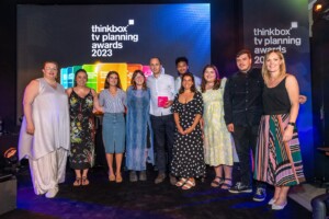 The Thinkbox TV Awards held at One Marylebone Road, 12th July 2023Photography by Fergus Burnett A group of ten diverse individuals poses with an recognition award at the "thinkbox tv planning awards 2023" event, smiling in front of a stage with event branding.