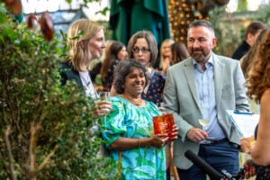 The Thinkbox TV Awards held at One Marylebone Road, 12th July 2023Photography by Fergus Burnett A diverse group of adults engaging in conversation outdoors at an event, with a woman in the foreground laughing and holding an recognition award, surrounded by plants and string lights.