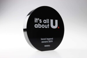 A black circular recognition award plaque reading "it's all about u", with the subtext "local legend award 2019" and the "veezu" logo at the bottom.