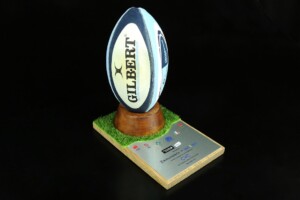 A rugby ball branded with "gilbert" displayed on a wooden stand with a grassy platform, against a black background. the stand includes various logos.