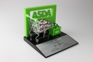 A deal toys model display featuring a miniature shopping cart filled with groceries beside a green asda basket in front of a large asda logo, presented on a base with the walmart and asda logos.