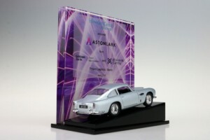 A model of a silver Aston Martin db5 displayed on a black stand, with a purple backdrop featuring multiple deal toys logos including "howden group" and "astonlark.