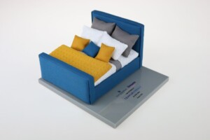 A miniature model of a bed with blue frame, yellow bedspread, and several gray and white pillows, displayed on a gray platform with a deal toys label.