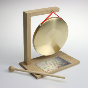 A small tabletop deal toys gong on a wooden stand with a mallet. there is a red ribbon attached to the gong and a plaque with text on the base. the design is simple yet elegant.