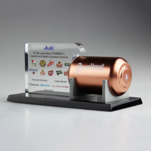 Deal toys featuring a metallic plaque with text and logos, accompanied by a sculpted golden beer can on a sleek black base, commemorating a business acquisition in march 2017.