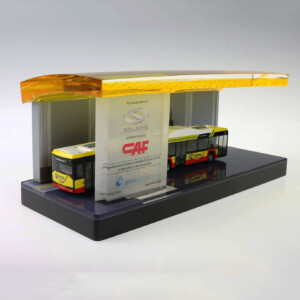 A deal toys model of a solaris bus stop with two buses, featuring a glass roof and promotional banners on the side, displayed on a gray platform.