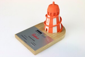 A 3d printed orange and white striped lighthouse deal toys model mounted on a wooden base with an attached silver plaque displaying the name "baltic" and other details.