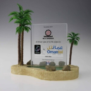 Transparent deal toys with text and logos, flanked by two miniature palm trees and set on a sand-like base with small rocks, commemorating a financial advisory role.