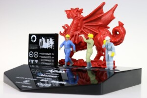 Miniature figures of workers in blue and khaki uniforms stand on a black platform, inspecting a red dragon statuette with outstretched wings, accompanied by a deal toys information card.