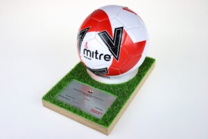 A red and white mitre soccer ball rests on a patch of green artificial grass set on a wooden base, which includes a silver deal toys plaque.