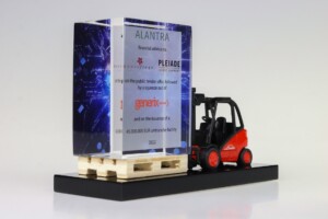 Model forklift with a linde logo carries a transparent deal toys tombstone with text and graphics, which sits atop a wooden pallet against a white background.