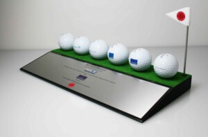 A display of six golf balls lined up on a green platform, each marked with a logo, next to a small golf flag, with a deal toys metallic plaque containing text in front.