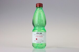 Transparent green deal toys plastic bottle with a red cap, labeled "aloe x ferrarelle botanic collection" in a white font on a gray background. the bottle features light water droplets on it.