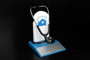 A stethoscope draped over a white, freestanding advertising deal toys display shaped like a shield with a blue cross, placed on a blue book titled "triton," against a black background.