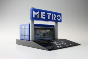 Detailed scale model deal toys of a metro station entrance with the word "metro" on a blue and white sign, displayed on a stand with clear lighting.