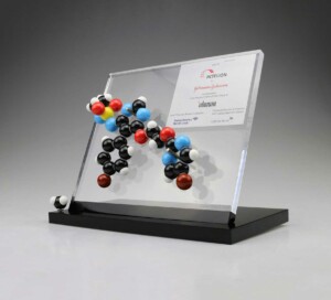 Clear acrylic deal toys with a 3d molecular structure model and a printed metal plate featuring logos and text, displayed on a black base.