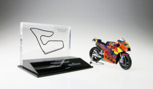 Deal toys with a model motorcycle next to a clear display with a track layout, both on a reflective surface. the motorcycle features a vibrant, sponsor-laden design.