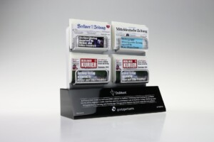 A deal toys display of miniature german newspapers in a black stand, featuring detailed front pages, each from different publishers like berliner zeitung and kurier. the background is light gray.