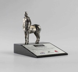 A silver deal toys statuette of a centaur, which is half man and half horse, mounted on a black base with a plaque that includes text and a couple of logos.