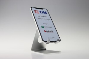 A smartphone displayed on a metal stand against a light background, showing a deal toys screen with the logos of tim, bnp paribas, and unicredit, dated october 2019.
