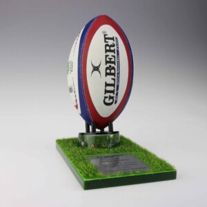 A gilbert rugby ball mounted upright on a green, grass-textured base with a silver deal toys nameplate, set against a soft gray background.