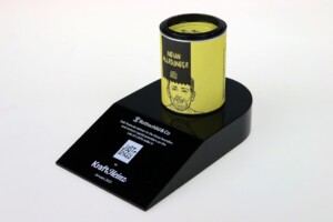 A tin of indian gunpowder on a black slanted deal toys stand with text and qr code. the tin features a yellow label and an illustration of a man's face.