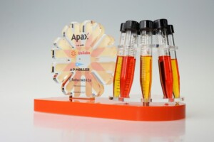 Transparent acrylic deal toys with corporate logos, next to a rack of test tubes containing colourful liquids, against a soft gray background.