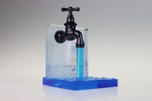 A transparent deal toys bottle filled with blue sanitizer has a black pump designed like an antique faucet, presented on a clear acrylic base with water droplets around.