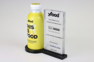 A yellow yfood bottle beside clear acrylic deal toys tombstone commemorating a strategic partnership with nestlé, dated march 2023. the award is displayed on a gray surface against a white background.