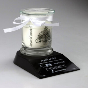 Deal toys candle labeled "fluffy cotton" on a black display stand with a logo and text detailing the brand. the candle is in a glass jar tied with a white ribbon, on a light grey background.