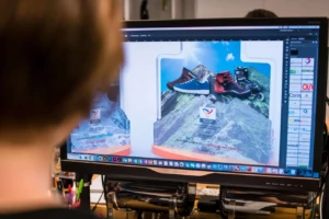 A financial tombstones designer views a computer screen displaying an deal toys design for hiking boots, with editing tools visible around the image.