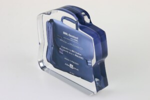 Transparent acrylic financial tombstones shaped like a briefcase with engraved text, possibly recognizing corporate or professional achievements, displayed against a white background.