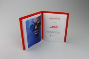 A red hardcover book opened to display a page with a striking image of a person wearing blue glasses and another page presenting the logo "ardian" and text details.