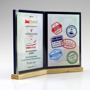 Clear acrylic financial tombstones displaying text and logos related to a corporate acquisition, adorned with colorful, embossed stamps that signify different stages of the transaction process, mounted on a wooden base.