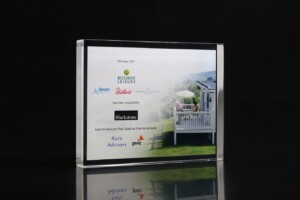 Transparent acrylic financial tombstones on a black background featuring a printed image of a porch overlooking a lush green landscape, with text announcing bourne leisure's acquisition by blackstone in february 2021.