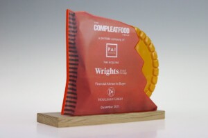 Financial tombstones on a wooden base, featuring a acrylic plaque with red and orange design, commemorating the acquisition of wrights by the compleat food group in december 2021.