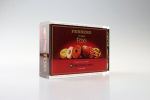 Clear acrylic financial tombstones containing a box of fox's biscuits, showcasing various biscuits visible through the transparent package, set against a plain light background.