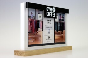 Financial tombstones for "gym + coffee" featuring a photo of the store interior with clothing racks and a customer, mounted on a wooden easel. the display includes text about a financial milestone.