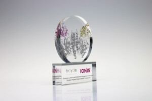 Clear acrylic financial tombstones with intricate silver and purple designs, mounted on a rectangular base that has the logos "ionis" and "biogen" along with text about oligonucleotide therapeutics.