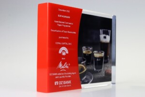 A red acrylic trophy with white inscriptions about a financial tombstones transaction involving coral capital dac and maltco, accompanied by a blurred background featuring glasses of dark beverage.