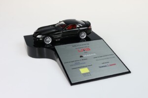 A black sports car model displayed on a tombstone with silver and red detailing. the plaque features several logos and text, indicating an award or recognition. the background is plain white.