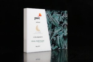 A two piece clear and white acrylic financial tombstone is positioned against a black background: one book is white with the pwc logo and text, and the other features an abstract green glass design.
