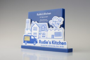 Financial tombstones for rudie’s kitchen, featuring a blue and white design with a food truck, buildings, and clouds, displaying the date "15th march 2021" and an address.