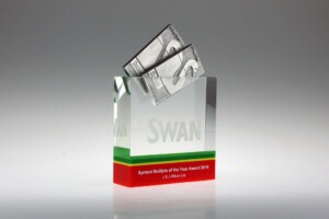 Clear acrylic financial tombstones with a green and red stripe at the bottom, displaying the text "giant swan, symbol multiple of the year award 2016, jj wilson ltd." a $100 bill origami swan is perched on top.